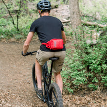 Load image into Gallery viewer, Person riding a bike in a forest with a red waist bag