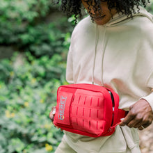 Load image into Gallery viewer, Person holding a red Meret bag outdoors with greenery in the background