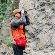 Load image into Gallery viewer, Person in orange vest and red bag using binoculars in a rocky outdoor setting
